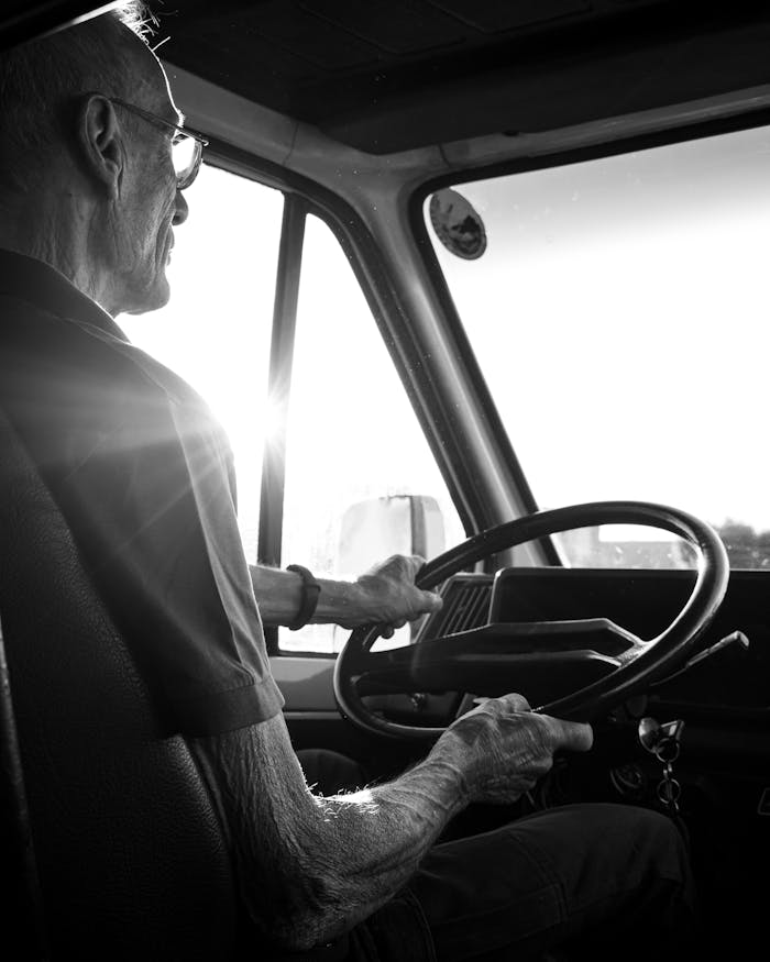 Elderly man driving a truck during sunset in black and white, capturing the essence of journey and experience.