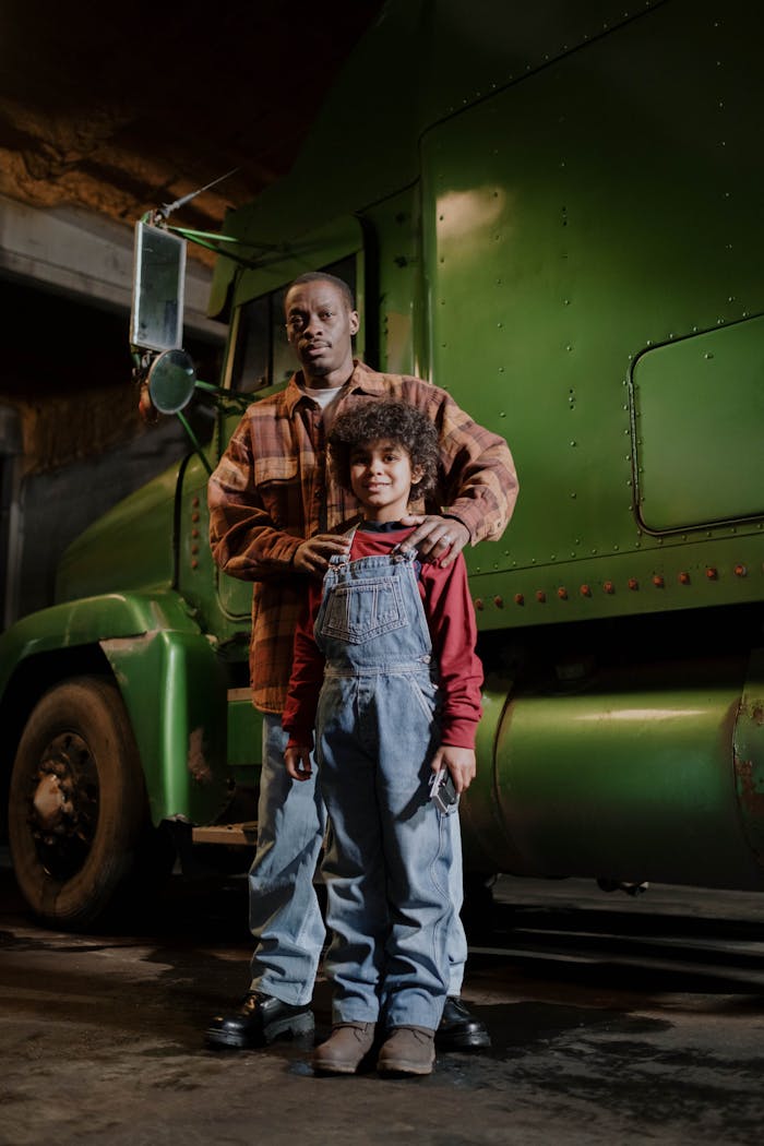 A father and son stand together in front of a large green truck in an industrial setting.