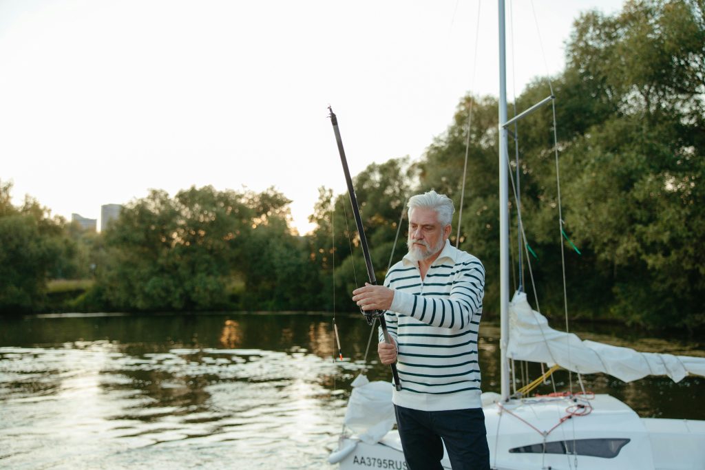 Elderly man adjusts his fishing rod aboard a sailboat during a tranquil outdoor setting.